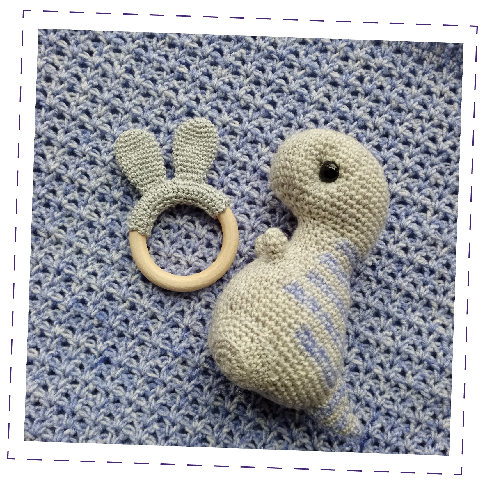 A handmade crochet blanket in a cool blue and grey mix, with a teether hoop with bunny ears and a soft toy dinosaur sitting atop it, in similar colours. 