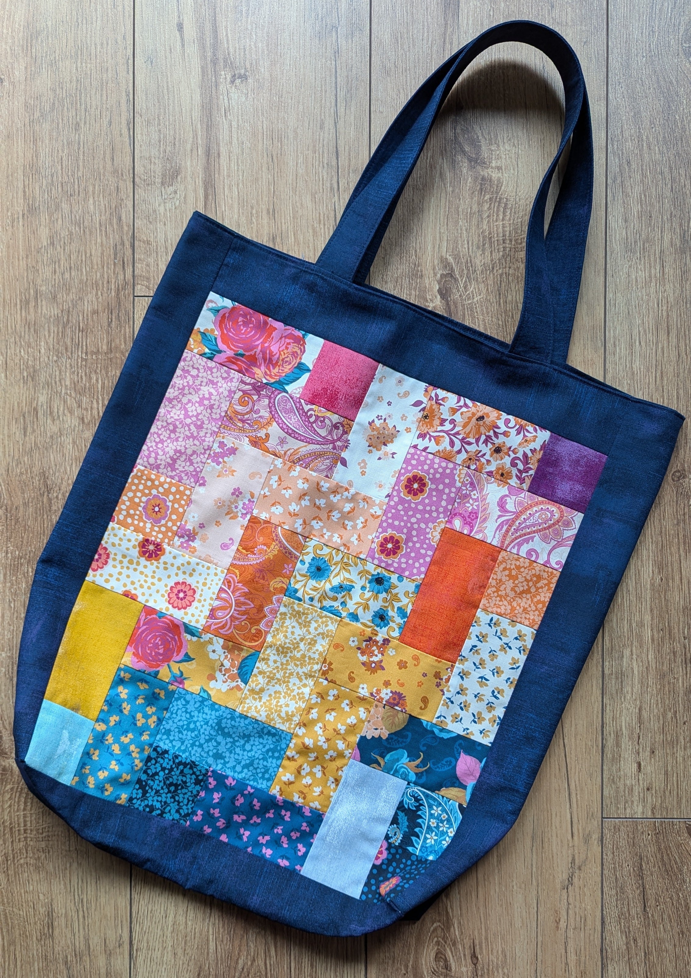 A handmade tote bag in a brick-like pattern featuring a variety of bright, colourful fabrics in pinks, blues and oranges, all framed in a deep blue fabric. 